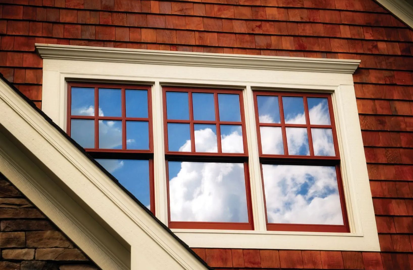 A triple-pane window with a white frame set against red shingle siding, reflecting a blue sky and white clouds.