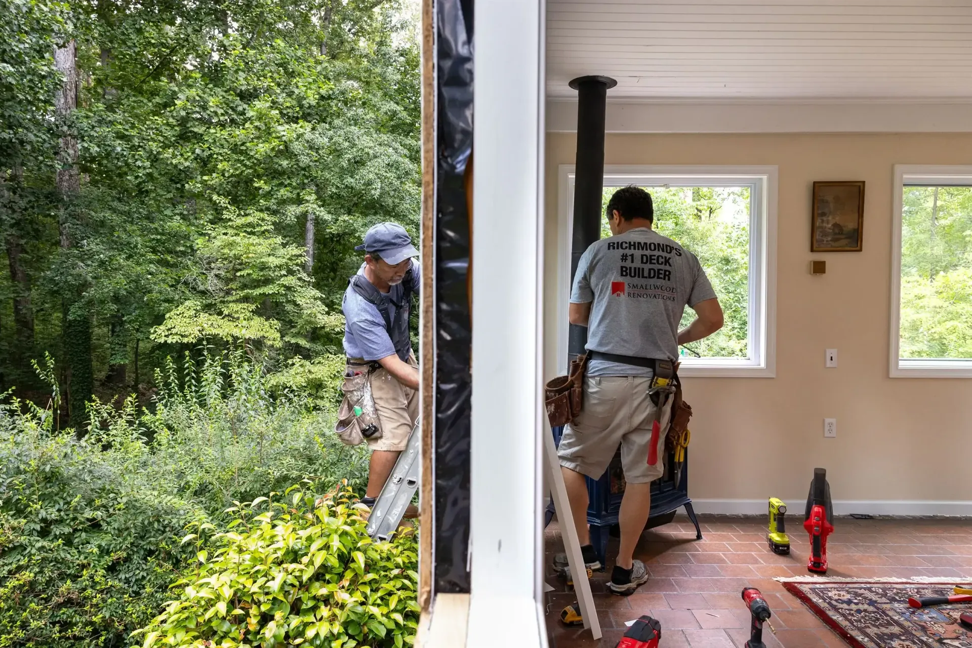 Two workers perform construction and interior renovations on a home, seen from an opening between an indoor and outdoor area.