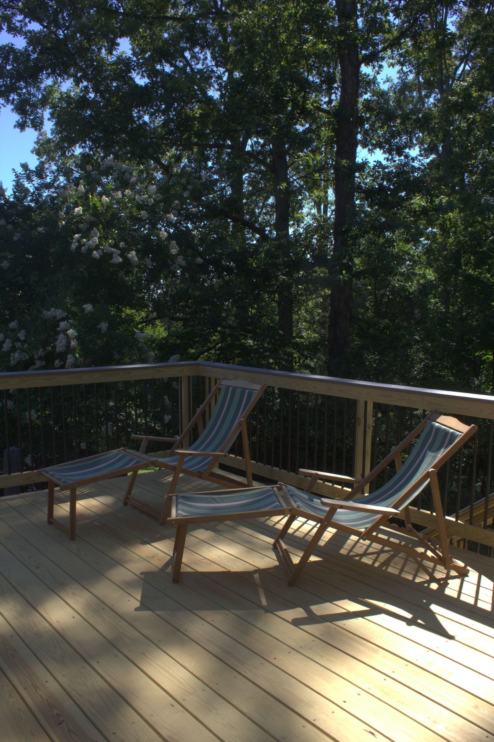A wooden backyard deck with a railing, featuring sliding glass doors leading to a grey house, surrounded by trees.