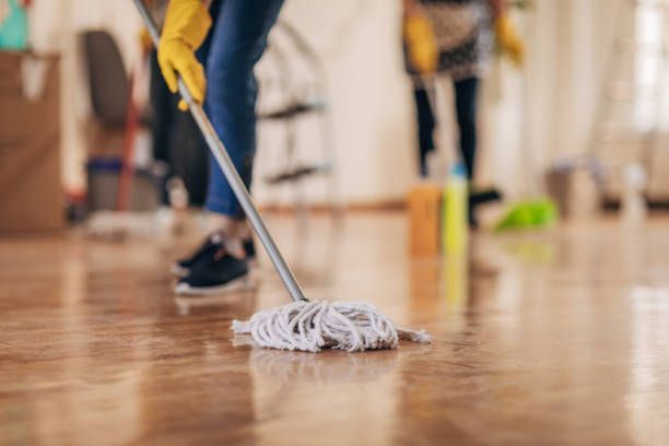 A person wearing yellow gloves mops a shiny wooden floor, with another person cleaning in the blurred background.