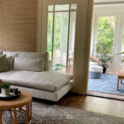 A sunlit living room features a white sectional sofa, a round woven coffee table, and a geometric rug on wooden floors.