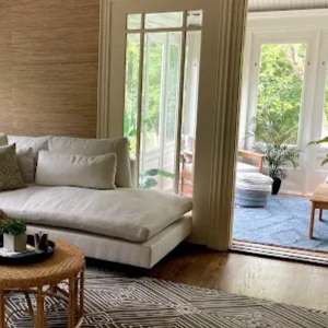 A sunlit living room features a white sectional sofa, a round woven coffee table, and a geometric rug on wooden floors.