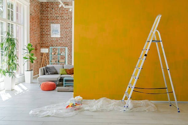 A yellow-painted wall with a ladder and drop cloth, set in a room with brick walls and a living area in the background.