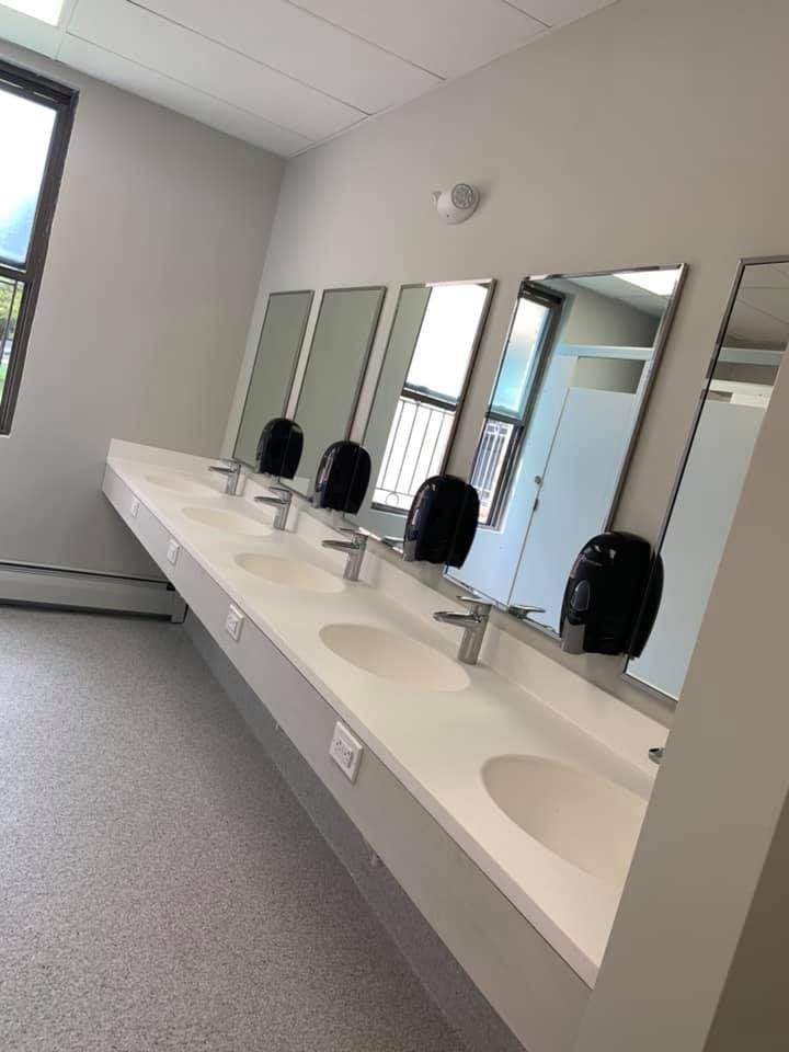A clean public restroom featuring a long white vanity with four sinks, silver faucets, and black wall-mounted dispensers.