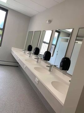 A clean public restroom featuring a long white vanity with four sinks, silver faucets, and black wall-mounted dispensers.
