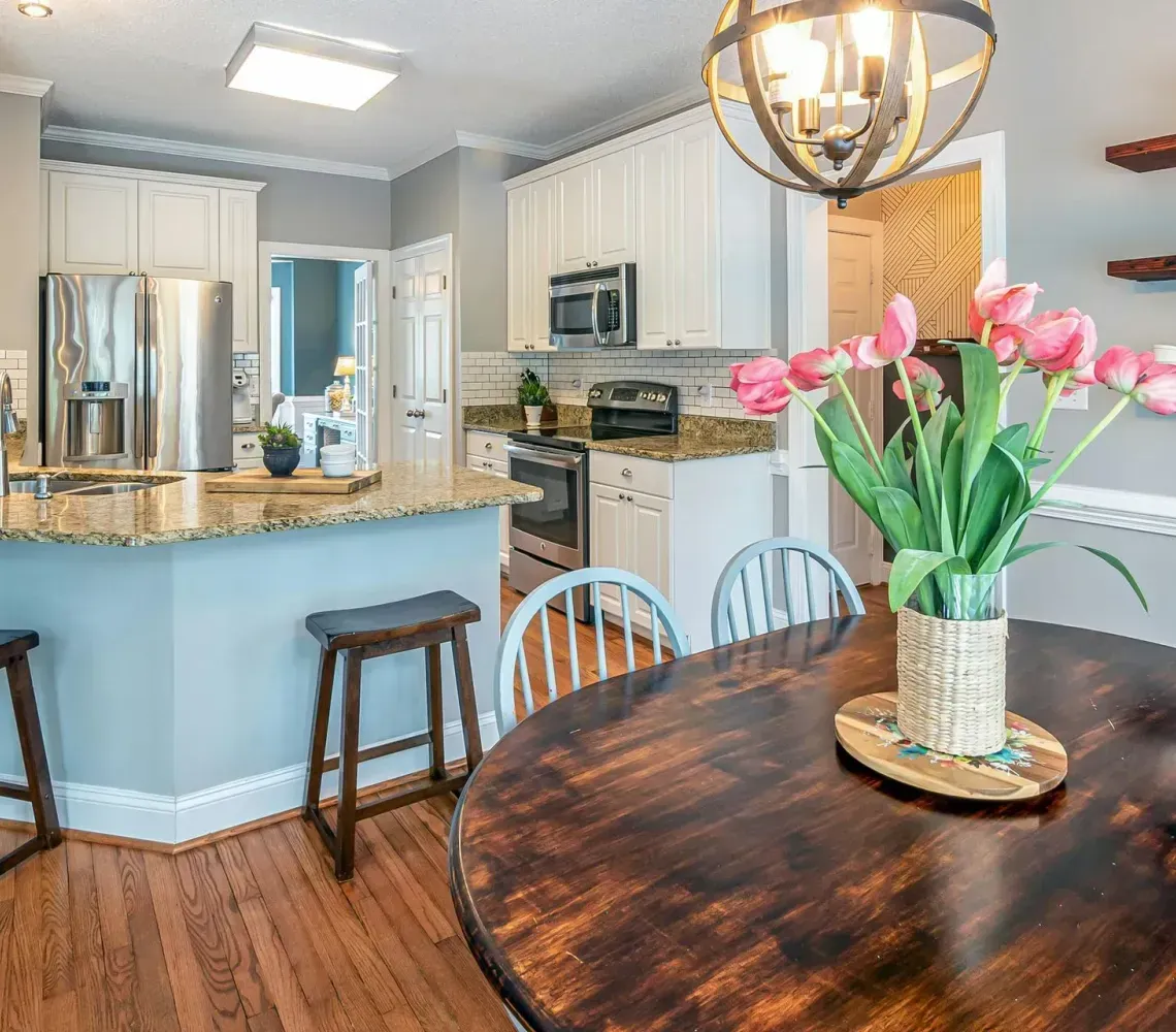 A bright kitchen with white cabinets, light blue island, two bar stools, wooden table, and pink tulips in a vase.