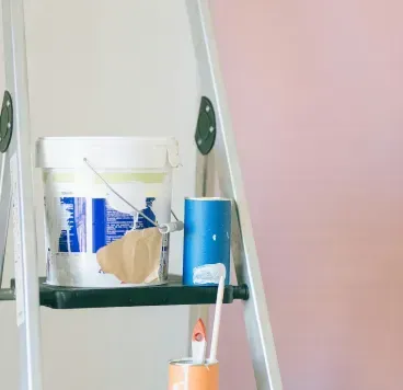 A silver step ladder holding a paint bucket, masking tape, an orange container with brushes, and paint brushes on a shelf.