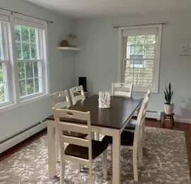 Dining room with a dark wooden table, four white chairs, light blue walls, and a patterned rug on a hardwood floor.
