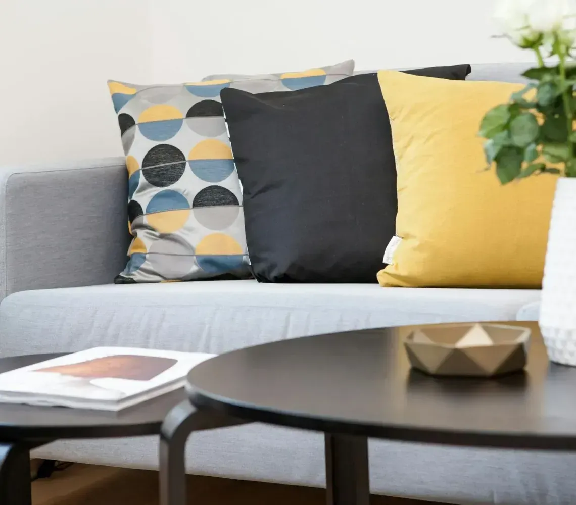 A gray couch with patterned, black, and yellow pillows beside a white vase of roses on a black coffee table.