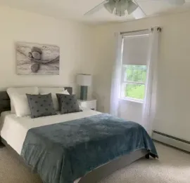 A bedroom with a bed featuring a gray throw blanket and pillows, a white wall with artwork, and a window with white drapes.