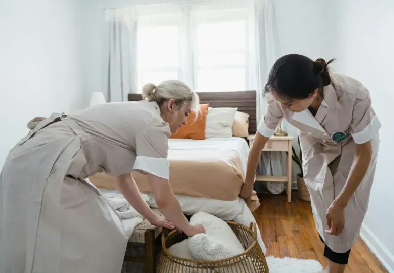 Two professionals in light-colored uniforms work together to prepare a bed in a bedroom.