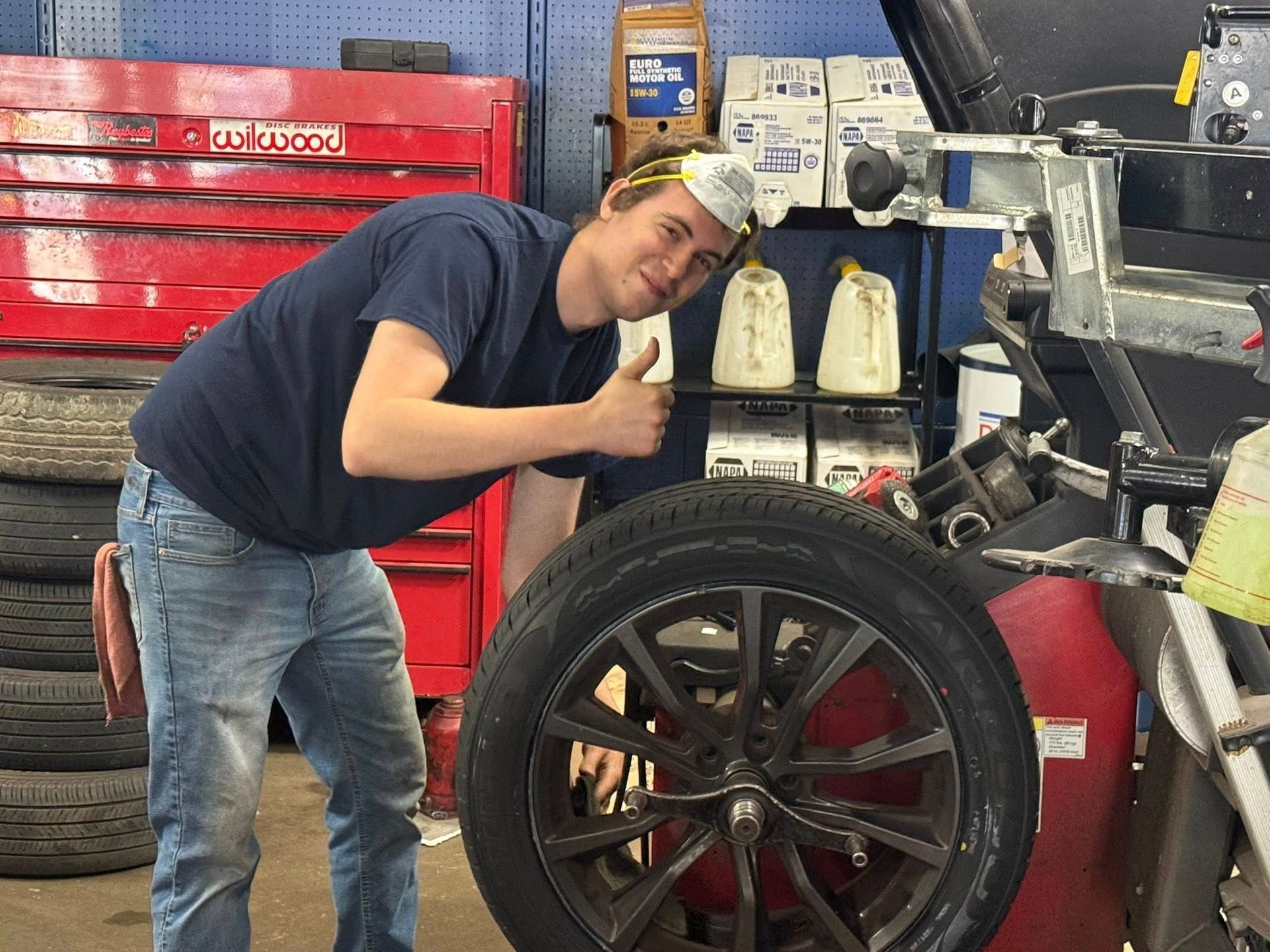 A man is giving a thumbs up while standing next to a tire in a garage.
