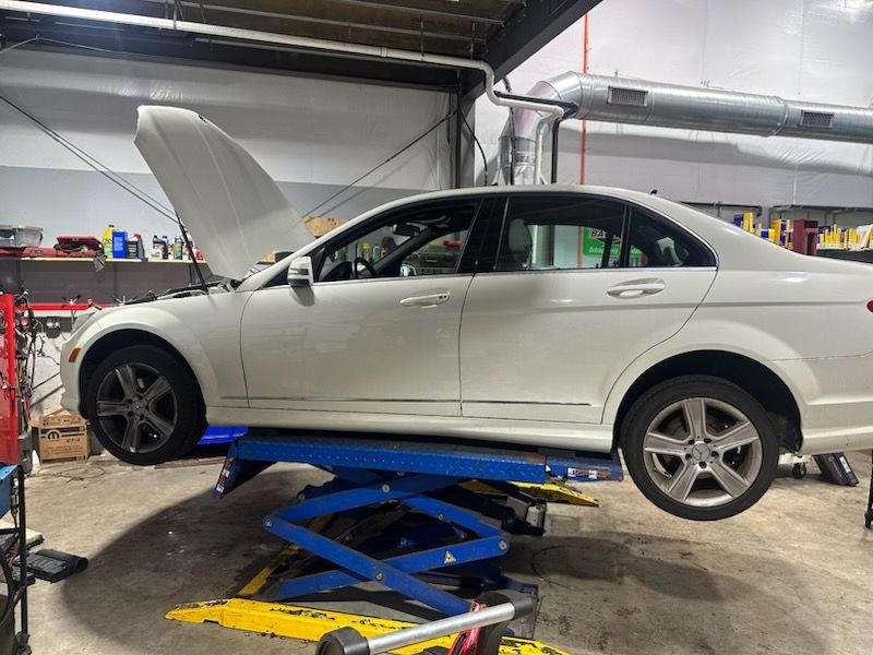 White Mercedes sedan on a blue lift with the hood open in an auto repair shop.