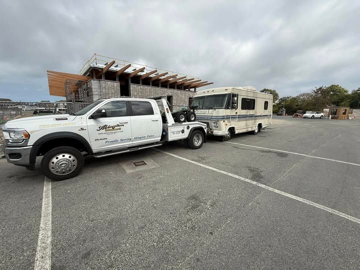 A white tow truck pulling an RV on a paved area near a building under construction. Cloudy sky.
