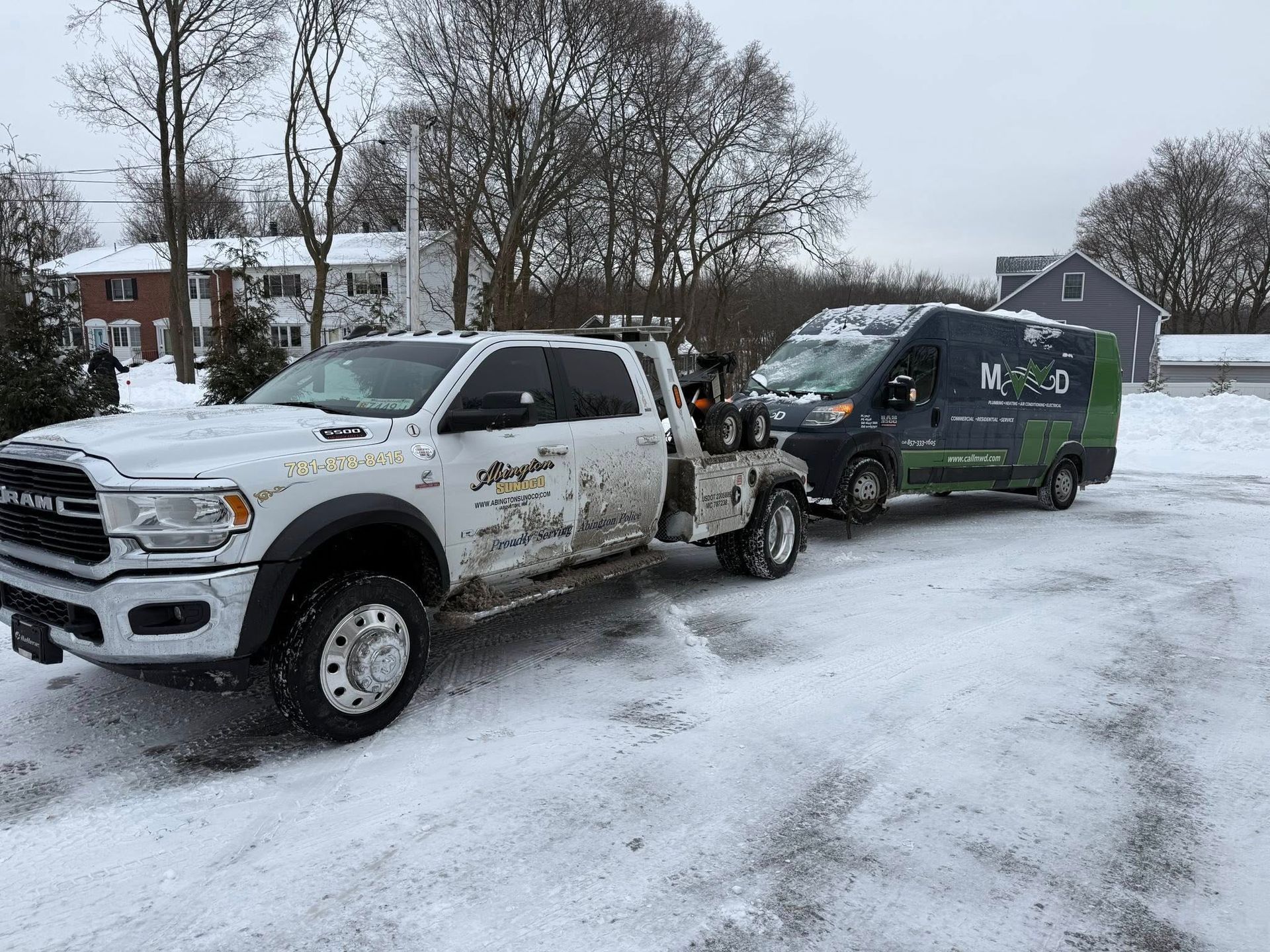 White tow truck towing a black van with green logo on snow-covered ground. Gray sky.