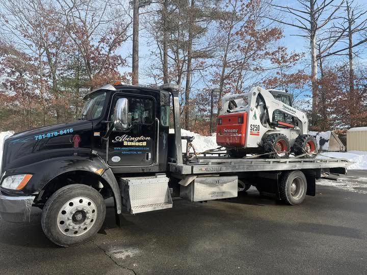 Black tow truck with a white and orange Bobcat skid-steer loader on its flatbed, outdoors on a snowy day.