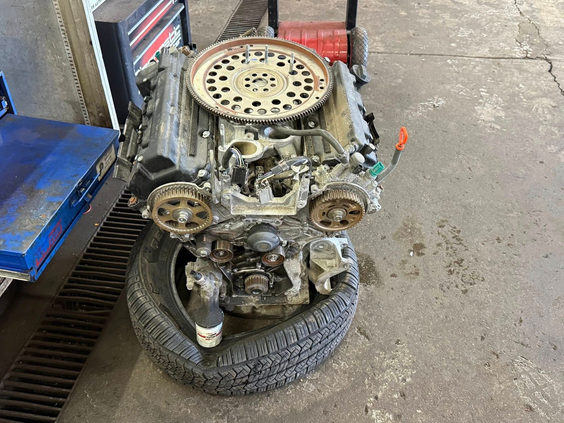 Engine resting on a tire in a garage, with various mechanical parts visible and a tool cart in the background.