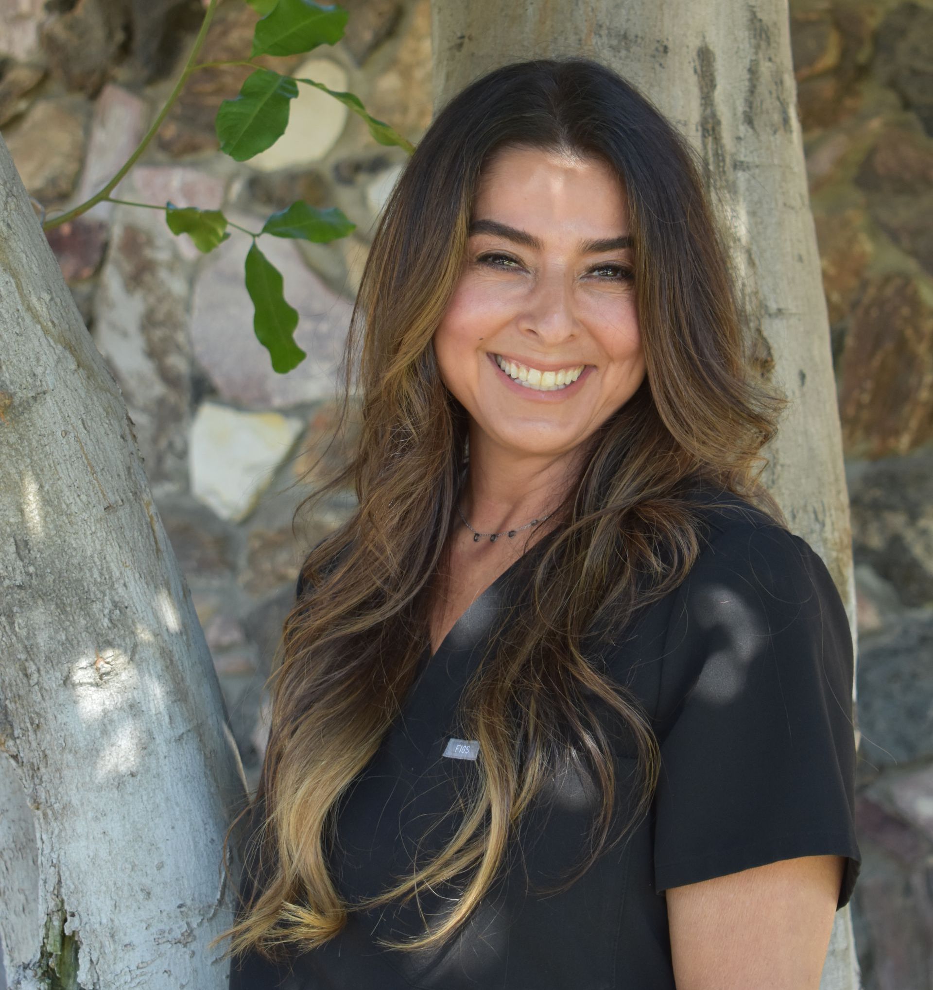 A woman in a black scrub top is smiling while leaning against a tree.