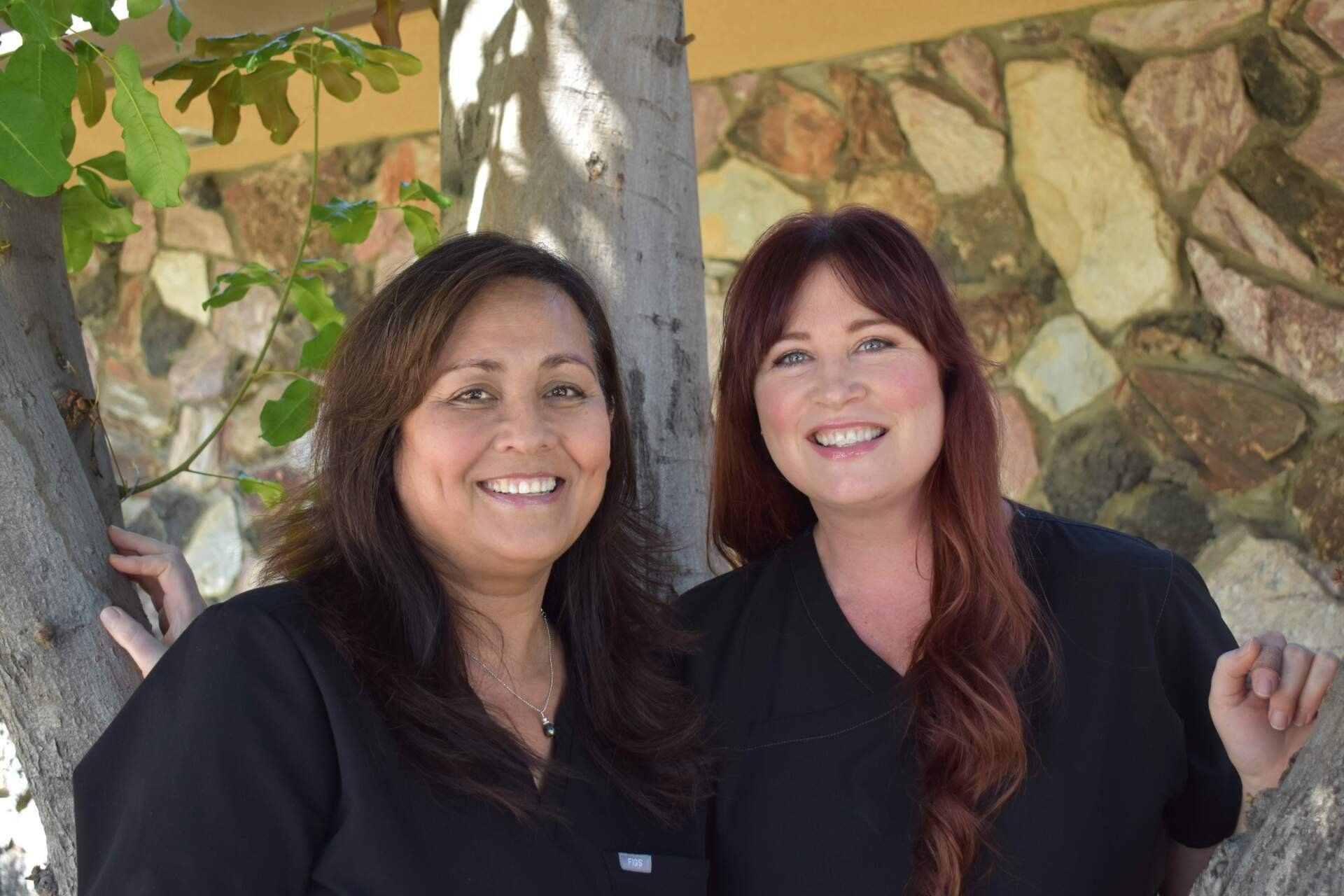 Two women are posing for a picture in front of a tree.