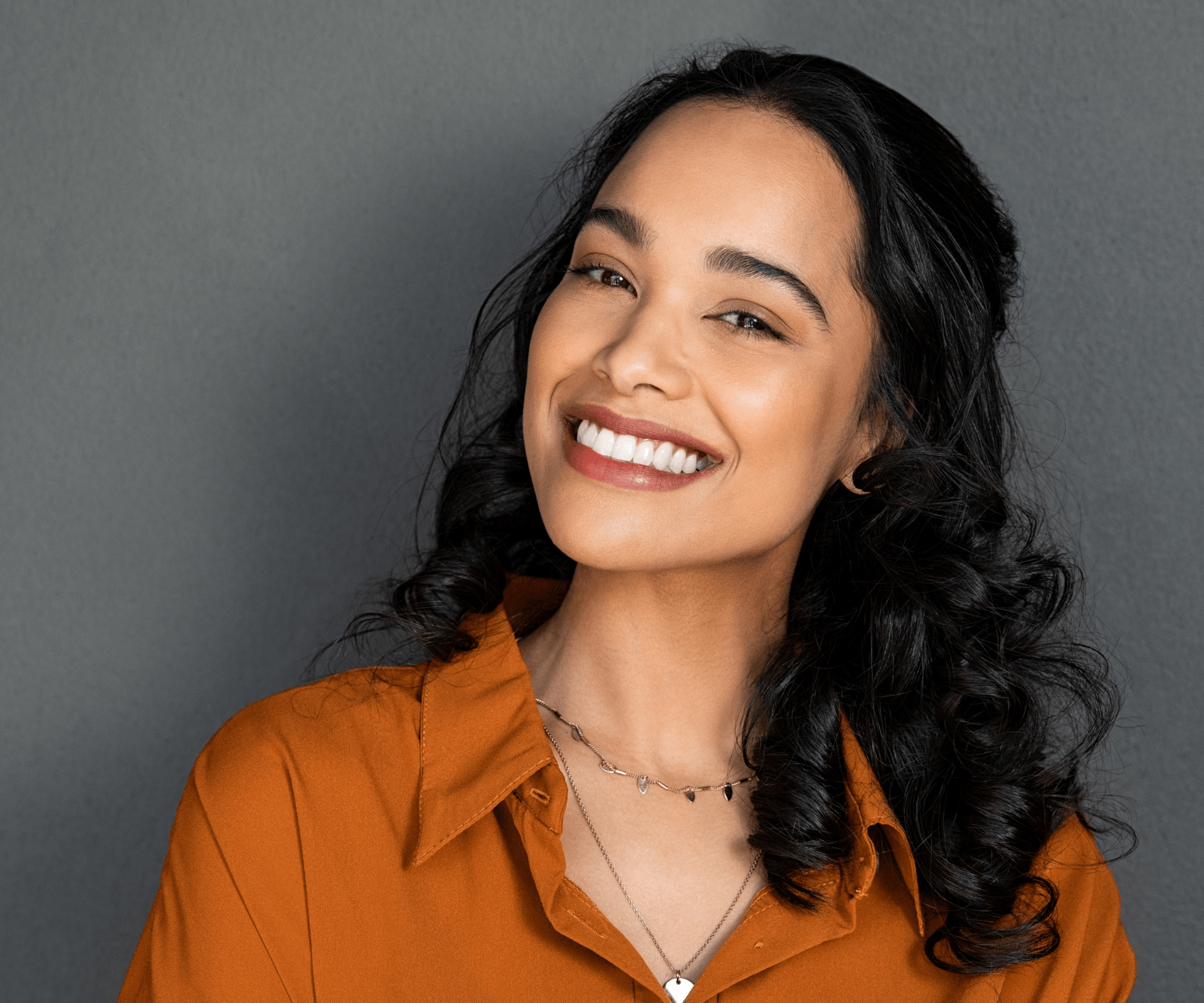 A woman wearing an orange shirt and a necklace is smiling.