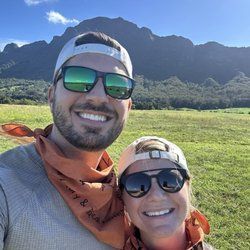 Two people smiling, wearing sunglasses and bandanas, in front of a mountain and green field.
