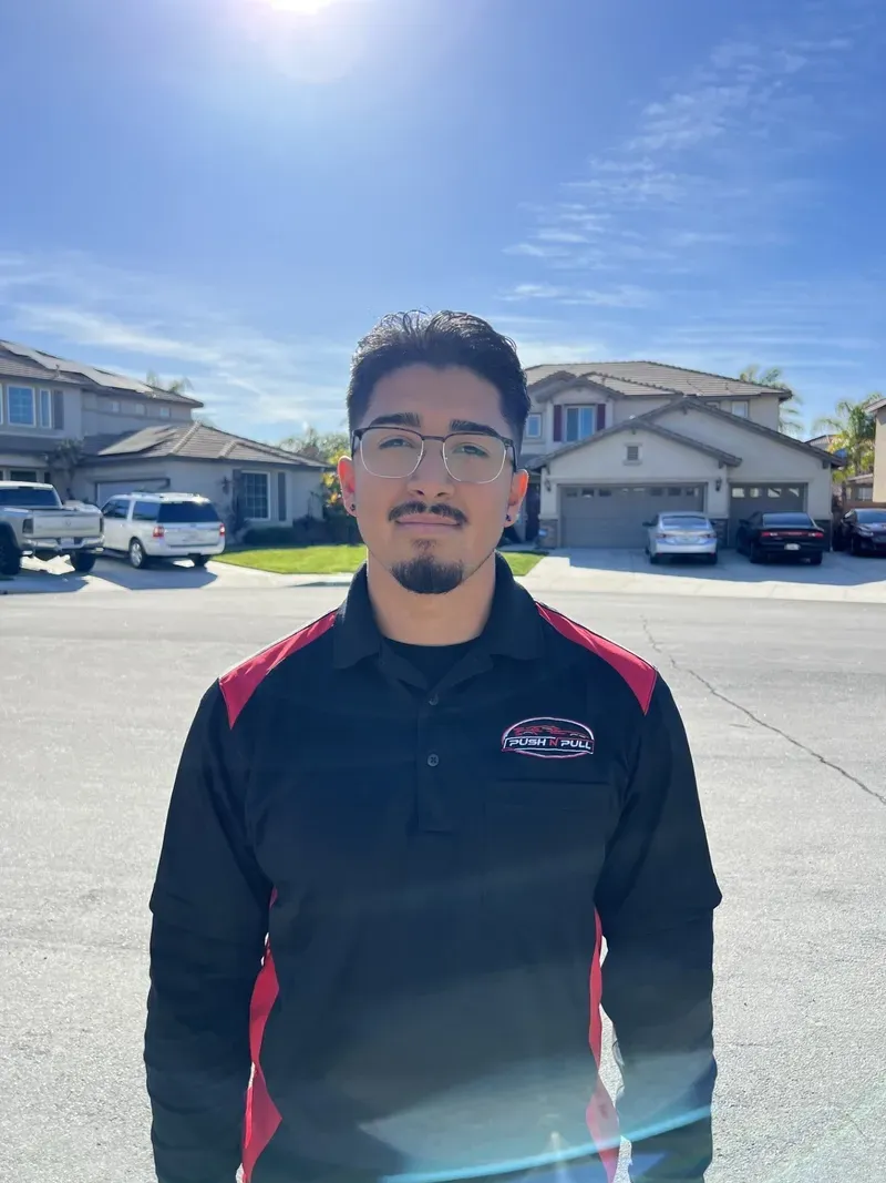 Man in black polo shirt with red trim, standing outdoors in front of houses on a sunny day.
