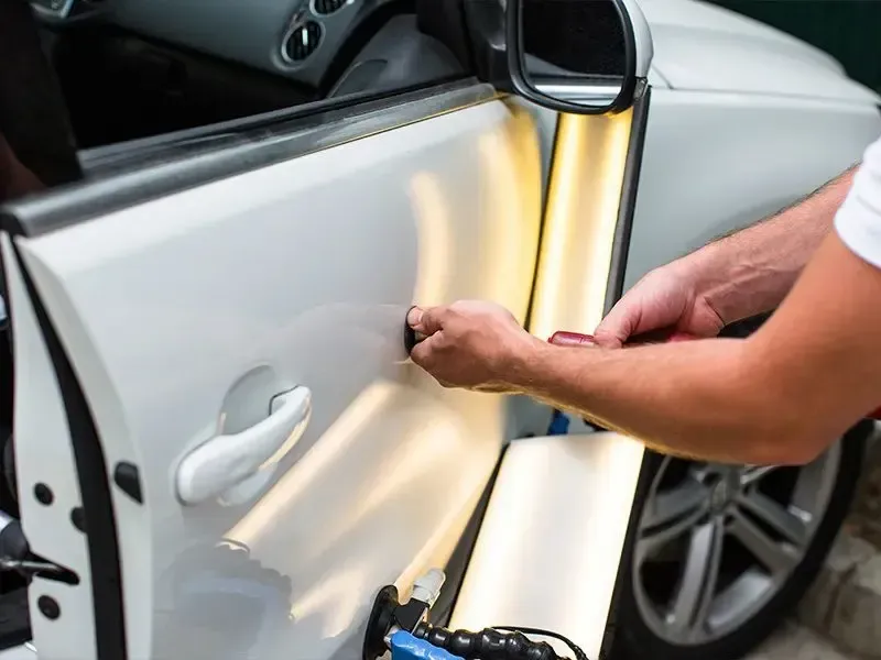 Mechanic in blue uniform inspects black car's fender, wearing gloves, in a garage.
