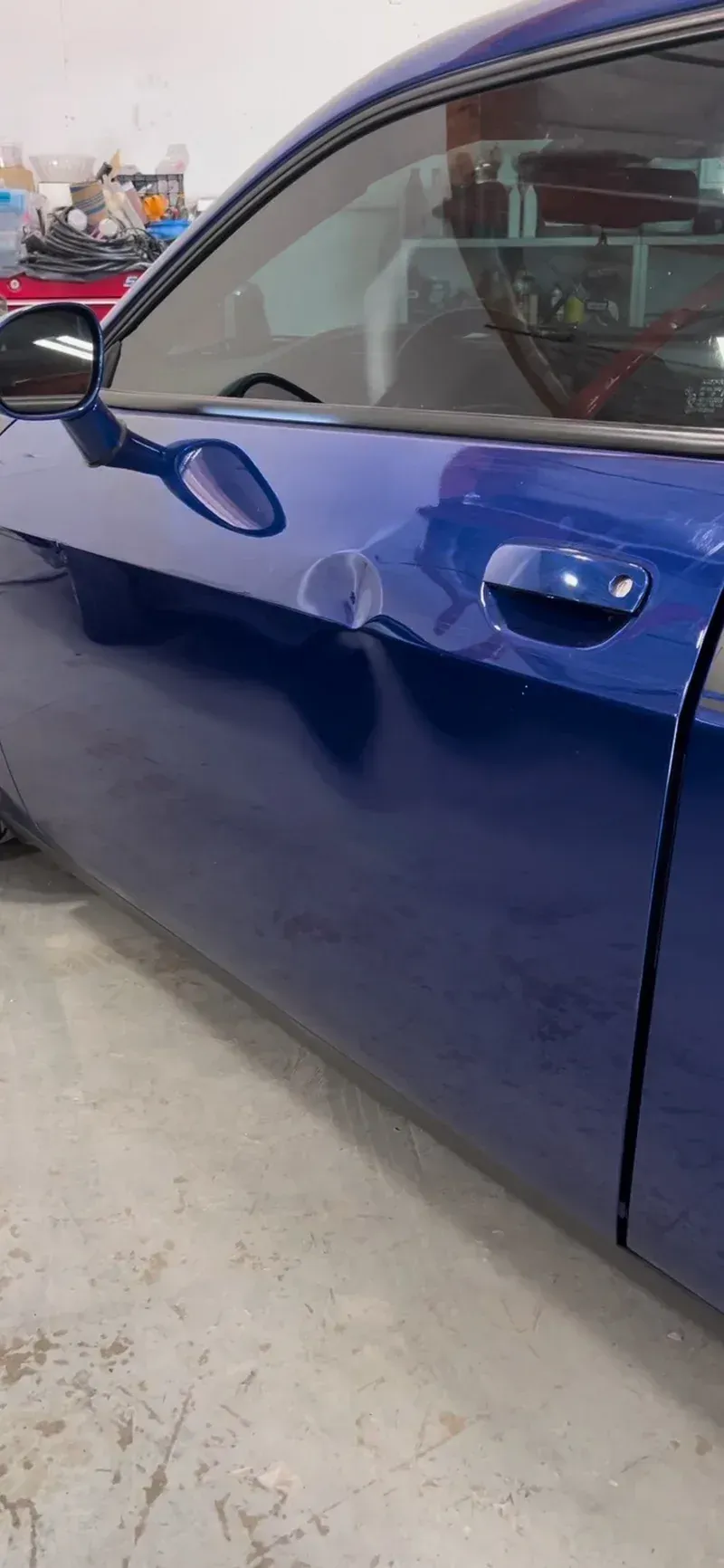 A damaged blue car door with a large dent is in a repair shop.