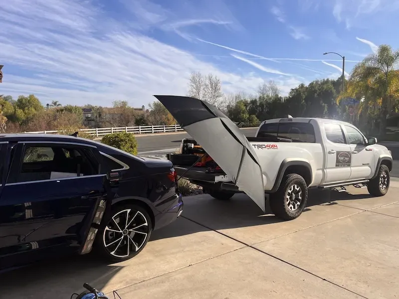 A white Toyota Tacoma truck with a black folding bed cover is parked next to a blue car.