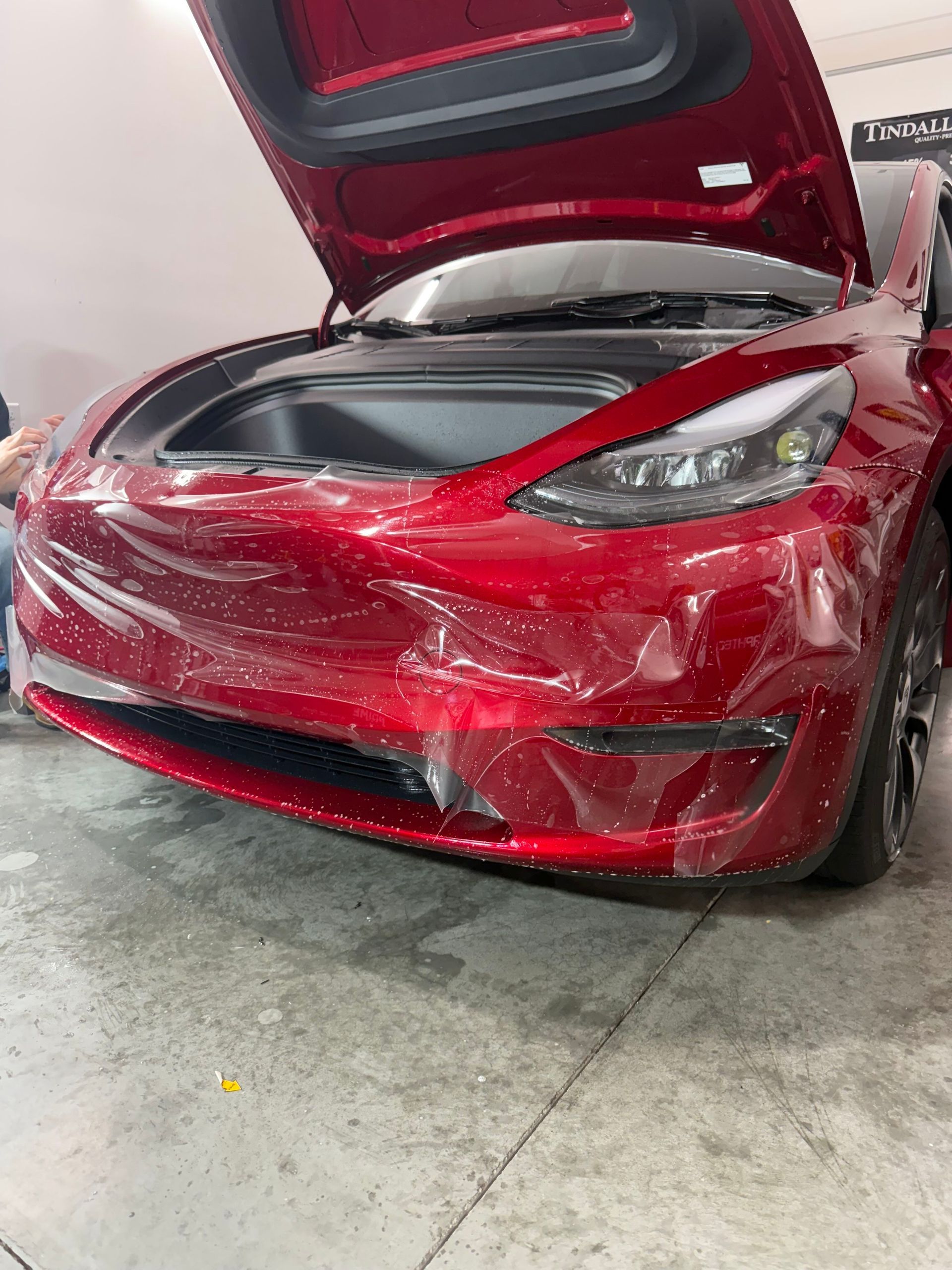 A red tesla model 3 with its hood open is wrapped in plastic.