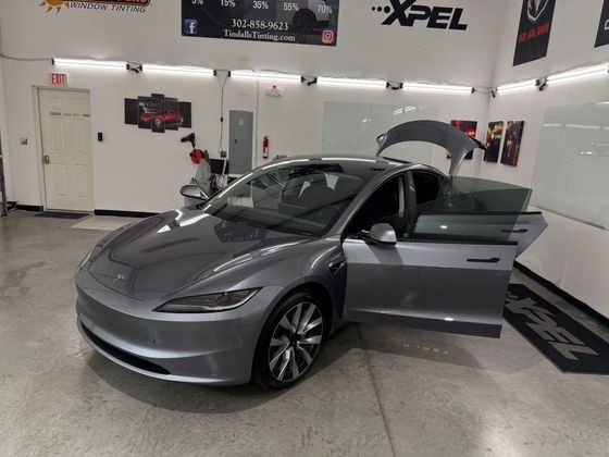 A gray tesla model 3 is parked in a garage with its doors open.