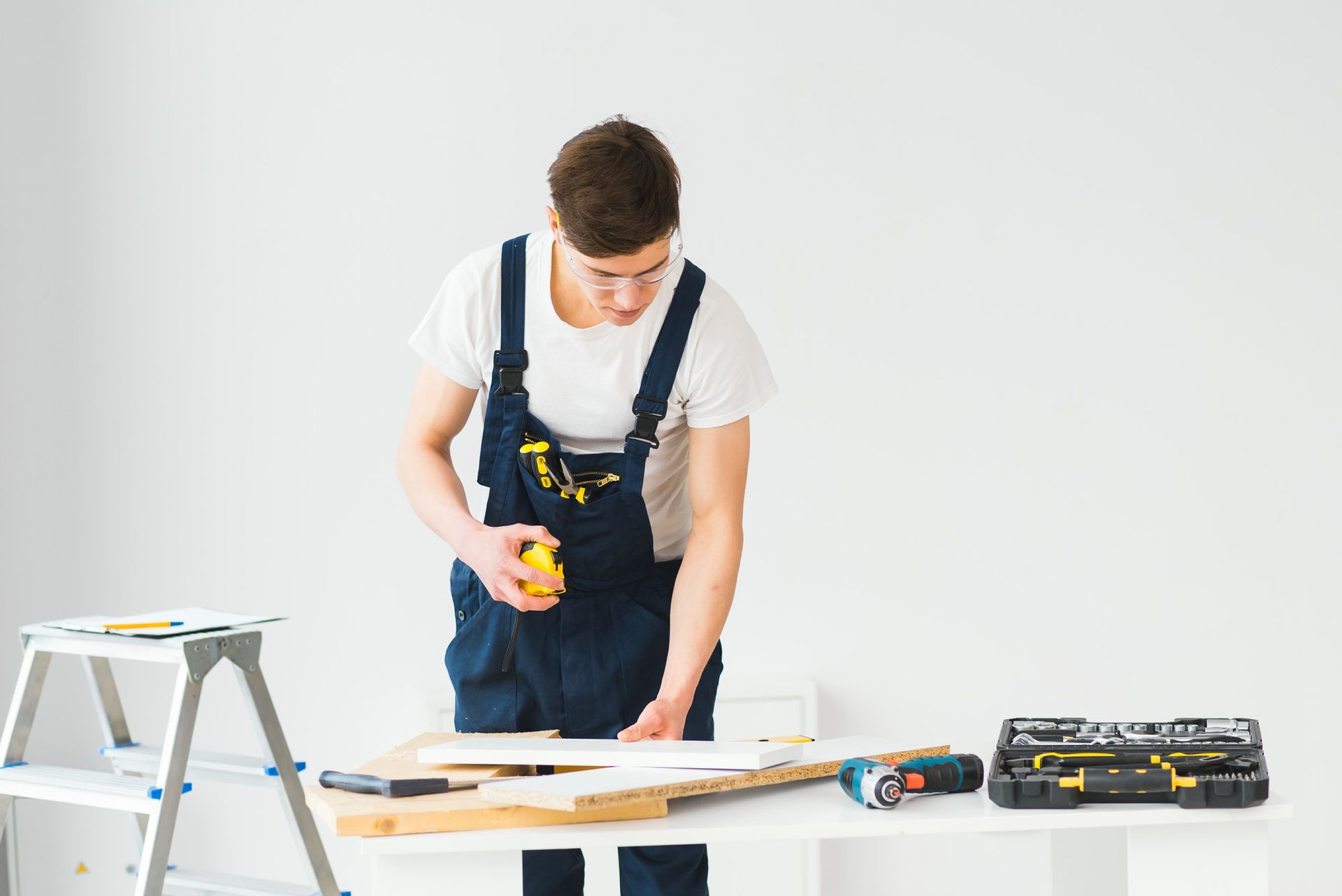 A person in blue overalls measures wood on a white table with tools, near a stepladder, against a white wall.