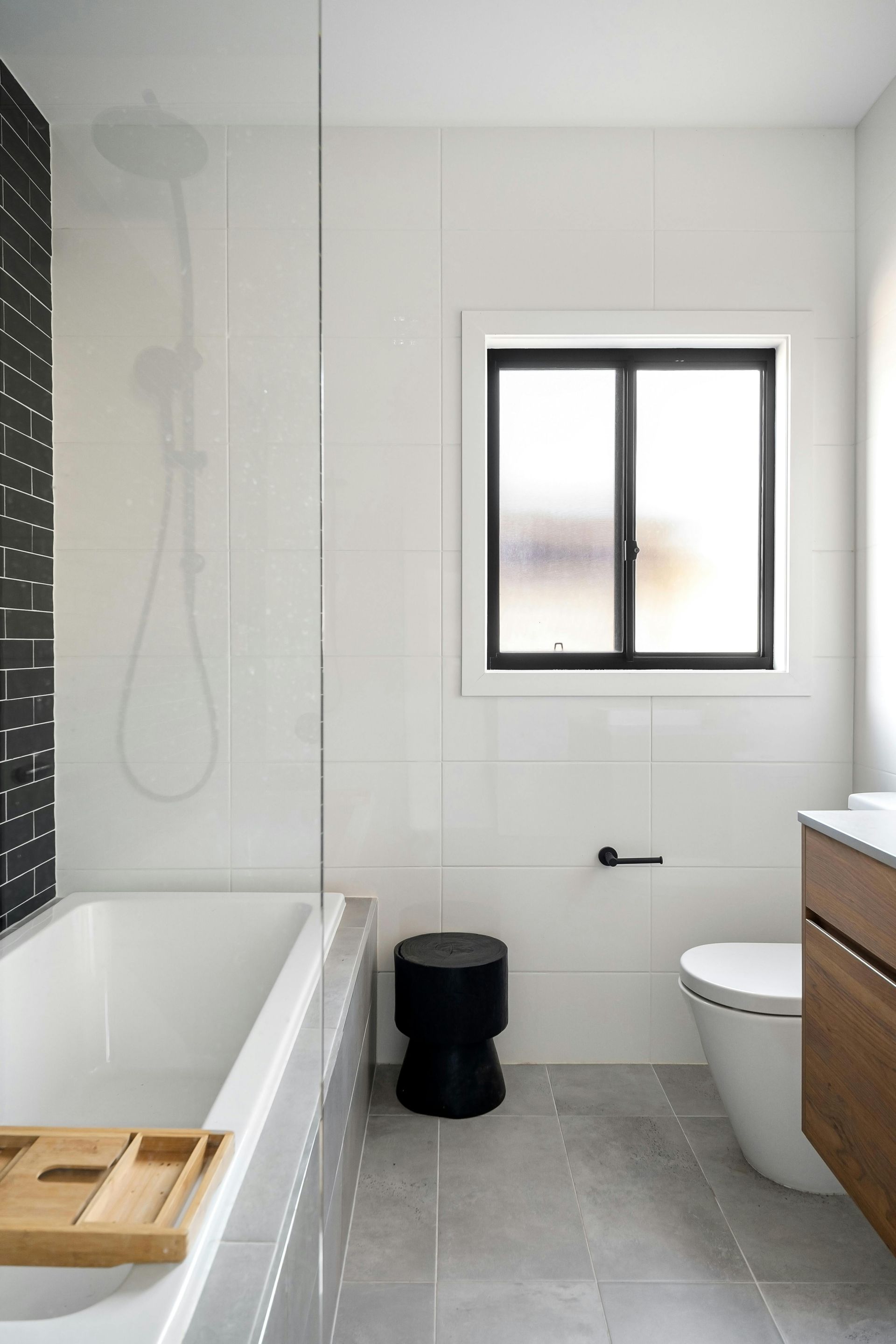 Modern bathroom: white tile, black accent wall, bathtub, toilet, window, wooden vanity, and black stool.