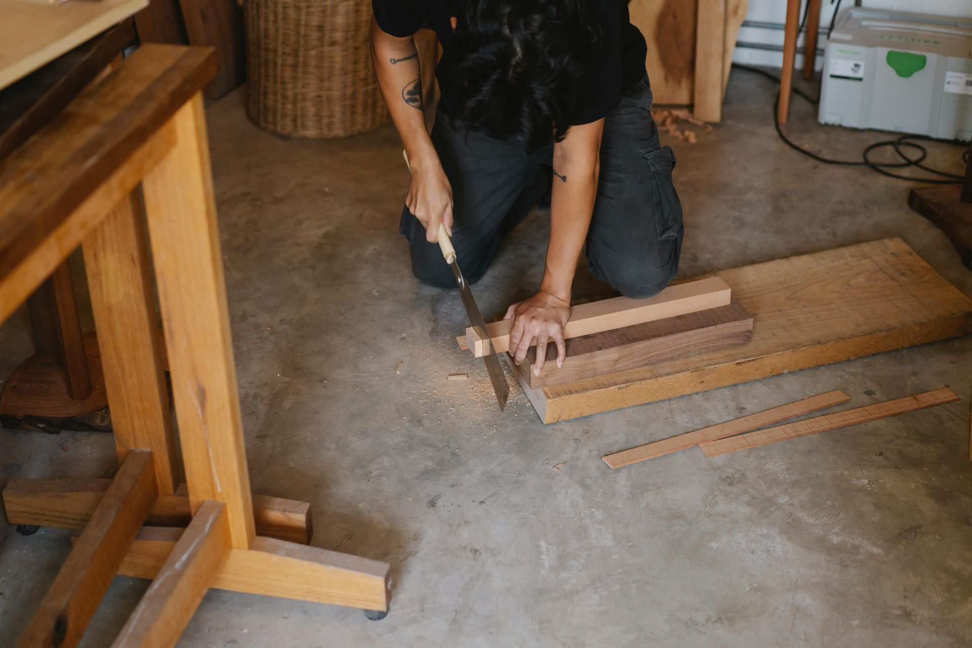 Person sawing wood pieces on a concrete floor in a workshop, near a workbench and materials.
