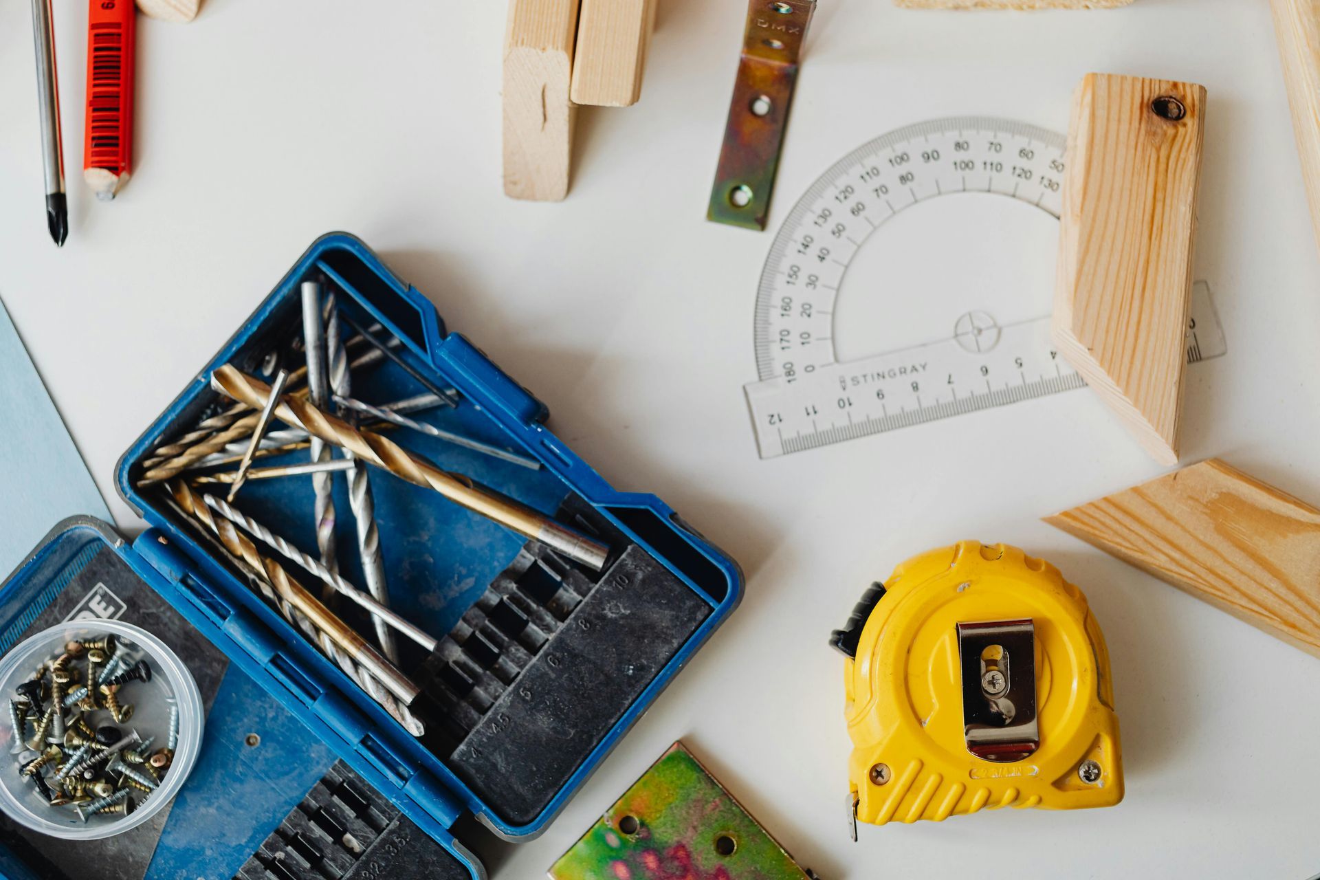 Workbench scene with tools: drill bits, screws, measuring tape, protractor, and wooden blocks, on a white surface.