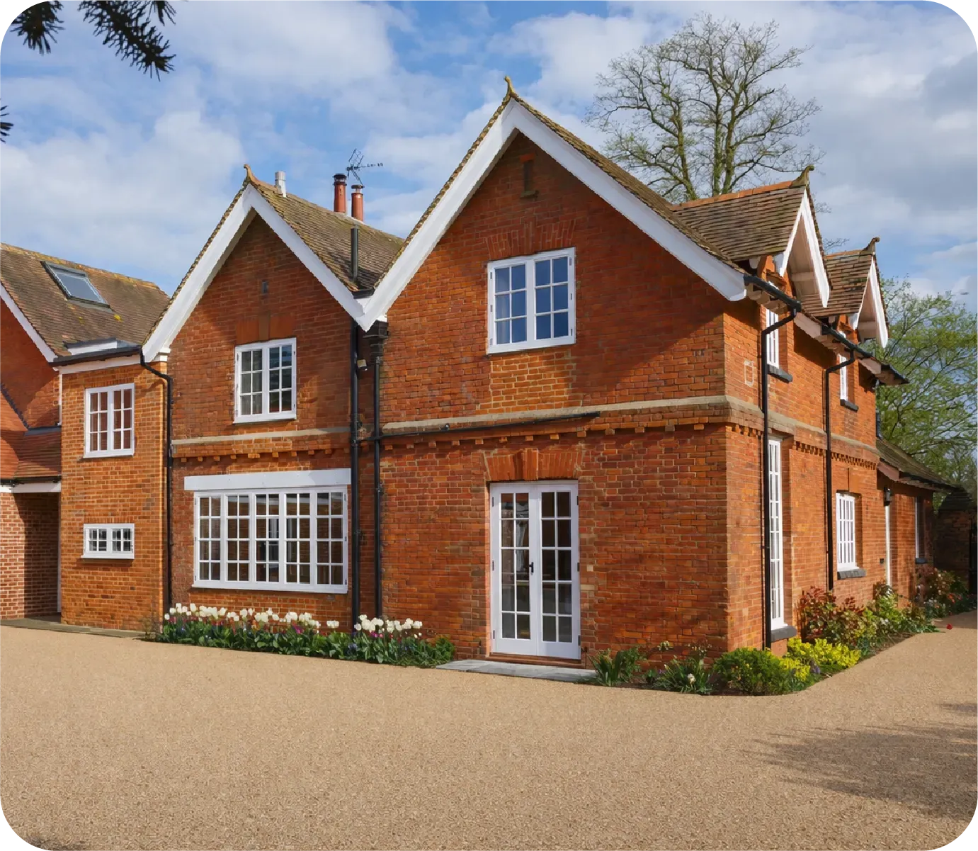 A two-story red brick house with white trim and a gravel driveway under a partly cloudy sky.