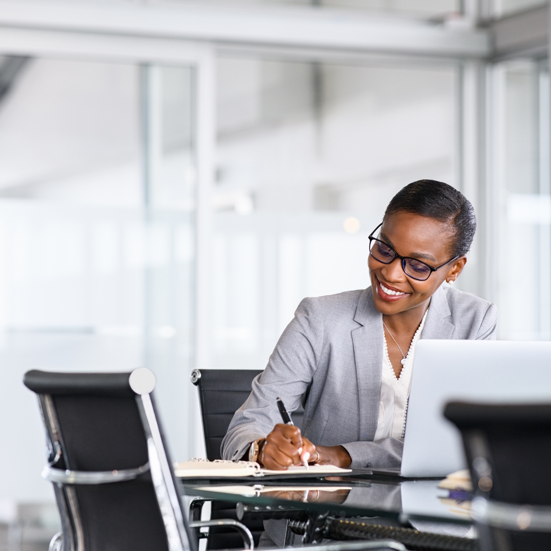 A woman is sitting at a table with a laptop and writing in a notebook.