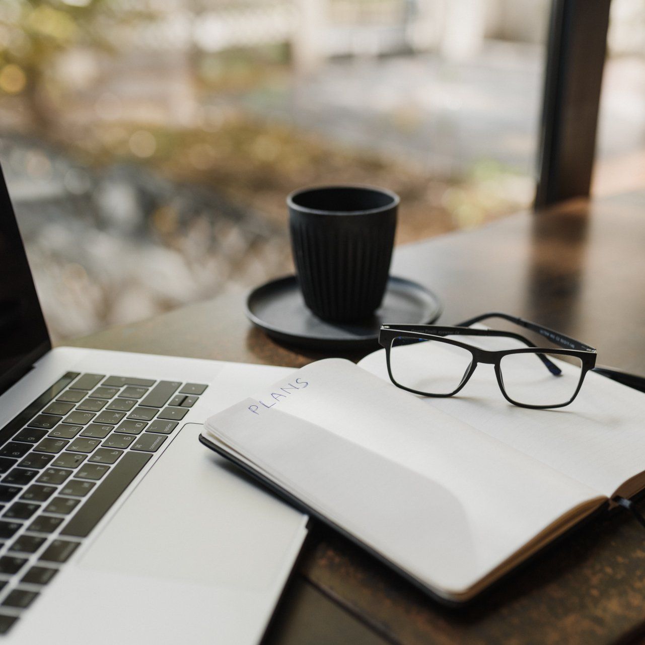 A pair of glasses sits on top of an open notebook next to a laptop.