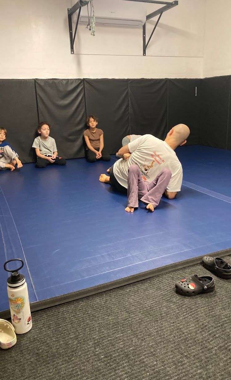 A man and a child practice Jiu-Jitsu on a blue mat. Several children watch from the side.