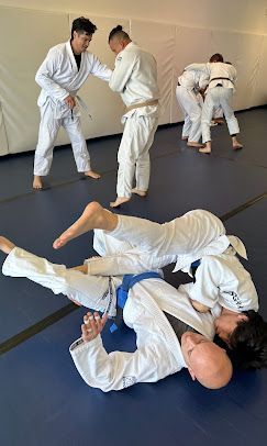 Judo class: students practice throws and ground techniques. A blue-belted man is pinned on the mat by another. Others spar.