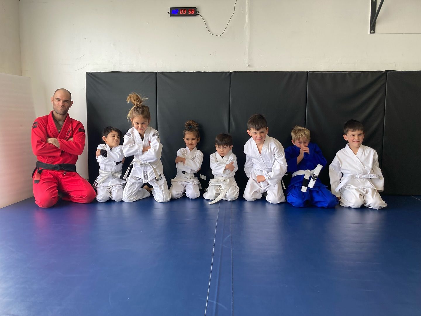 A group of children in karate uniforms kneeling with a martial arts instructor in a gym. The instructor wears a red gi; the children wear white.