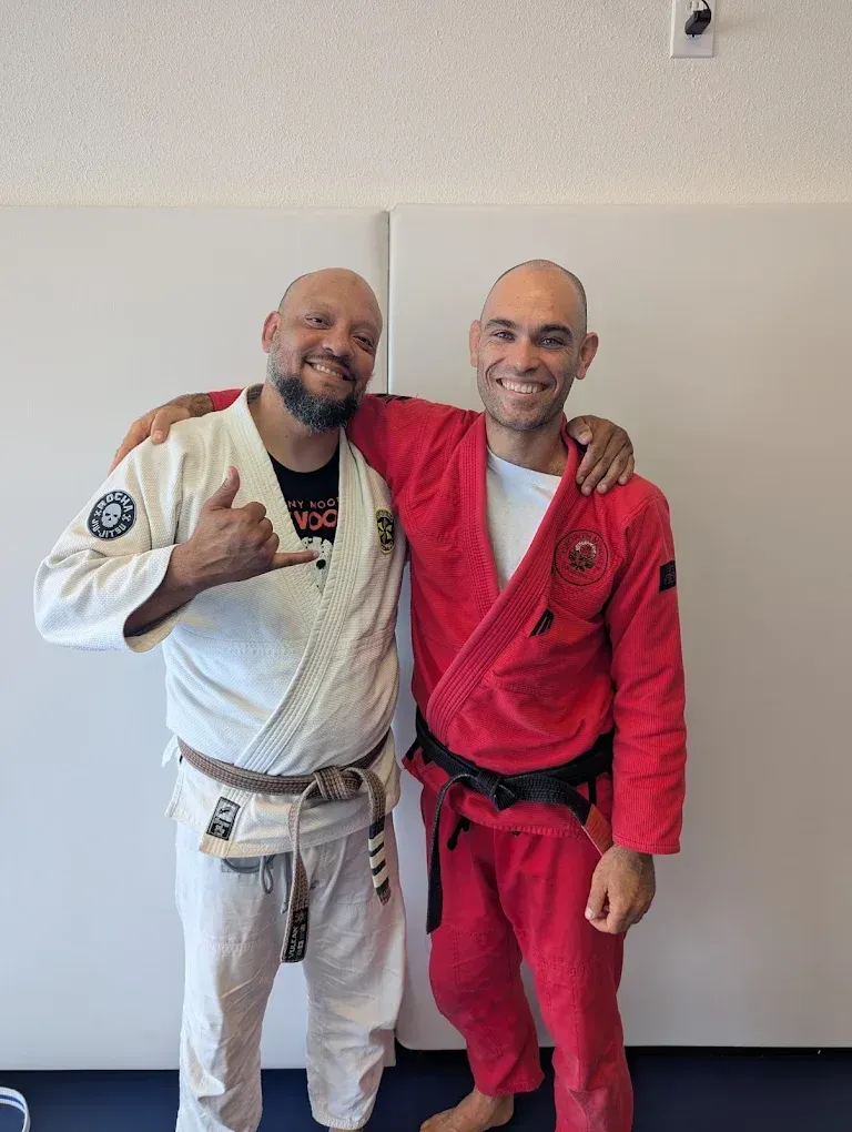 Two smiling men in martial arts uniforms pose arm-in-arm. One wears a white gi with a brown belt, the other a red gi with a black belt.