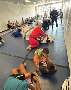 People practice Jiu-Jitsu on a blue mat in a gym. A red-clad instructor demonstrates moves as others watch and participate.
