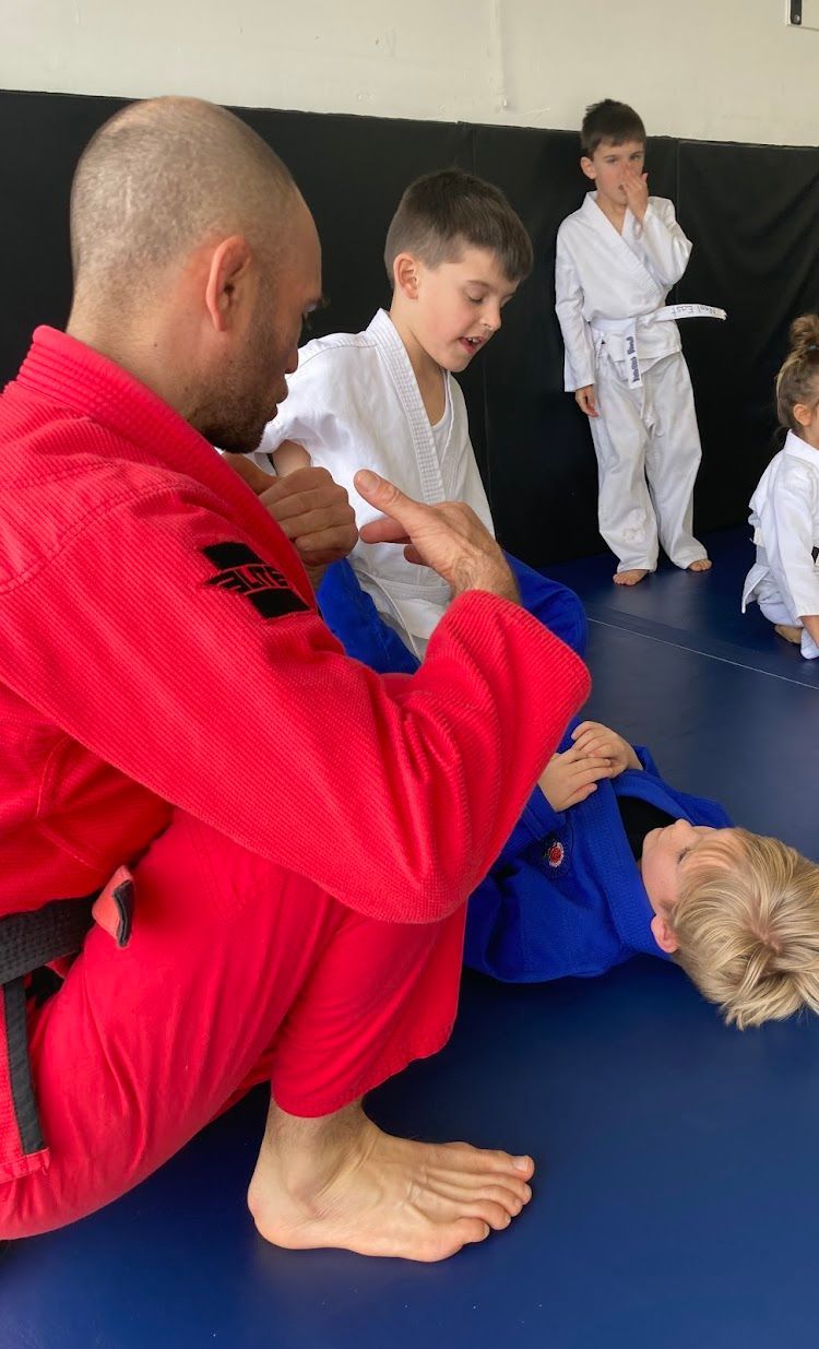 A bald instructor in a red gi demonstrates a technique to children in white gis on a blue mat.