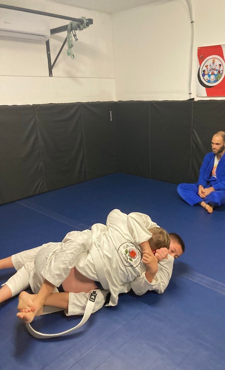 Two people in white gis practice a grappling technique on a blue mat in a gym. One person has the other in a headlock. A person in a blue gi watches.