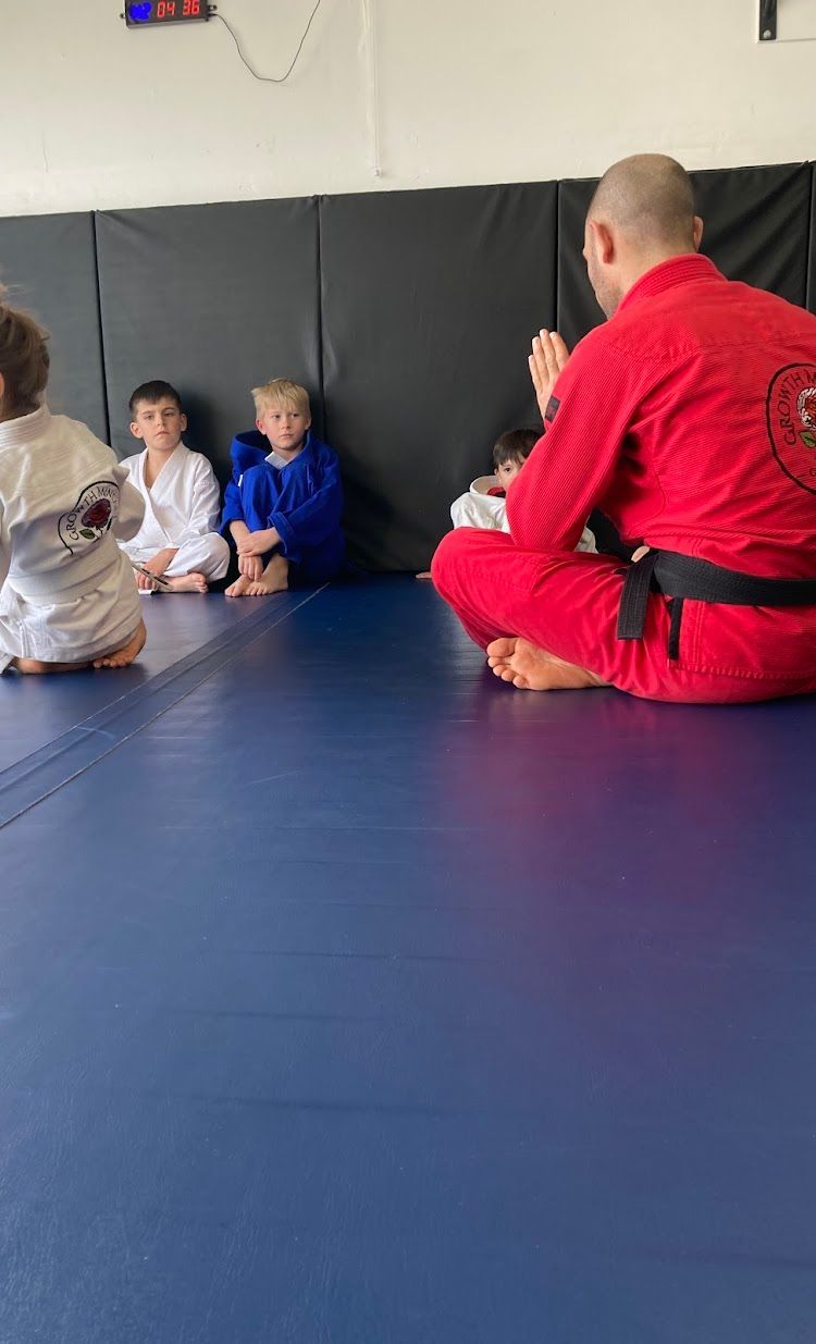 A martial arts instructor in a red uniform kneels, facing a group of children in training attire on a blue mat.