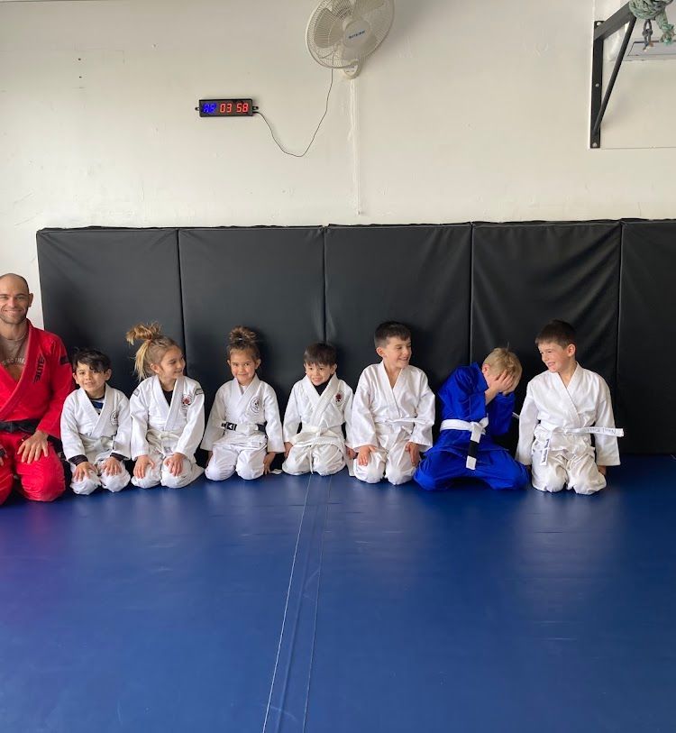 A group of children in white martial arts uniforms kneel next to an instructor in a red uniform on a blue mat.