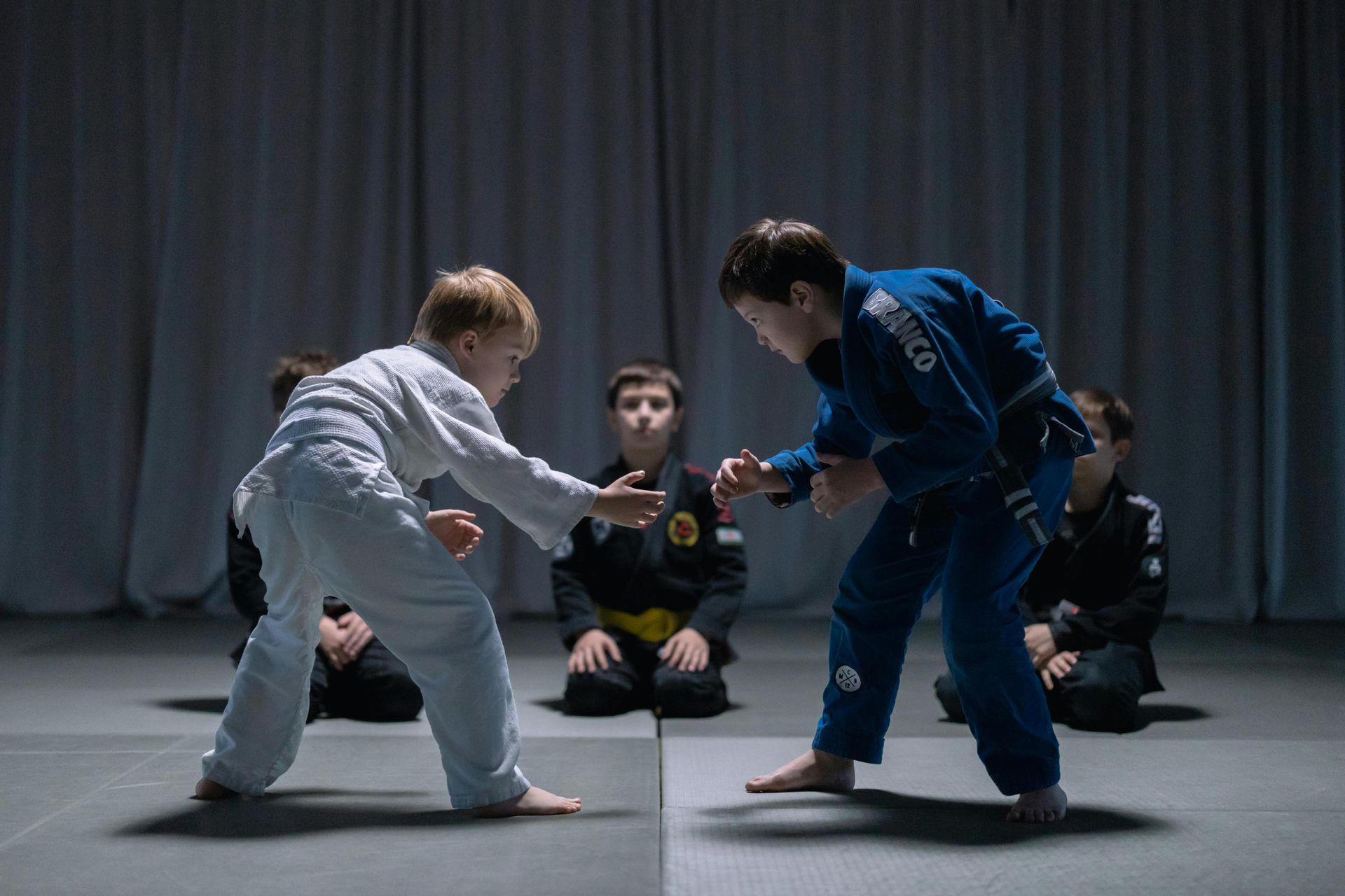 Two young boys in jiu-jitsu uniforms face each other on a mat. Other children watch in the background.