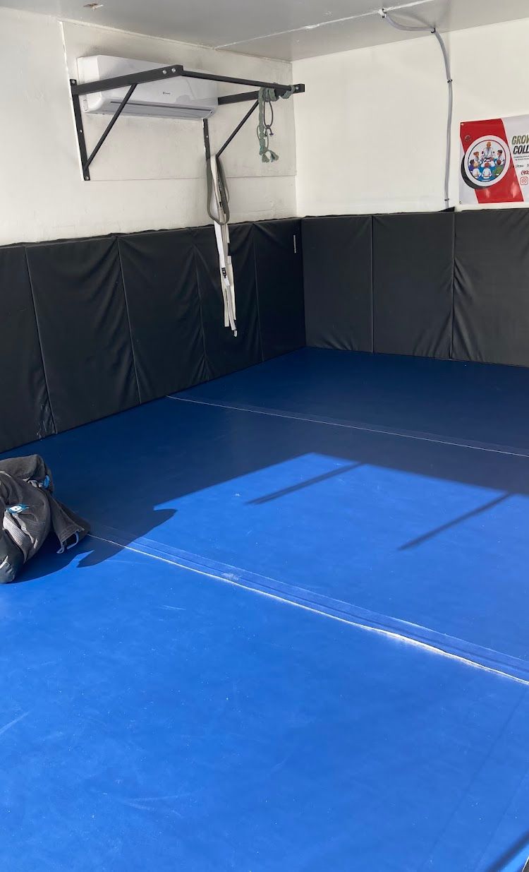 Blue gym mats cover a floor, with black wall padding and a pull-up bar attached to the wall.