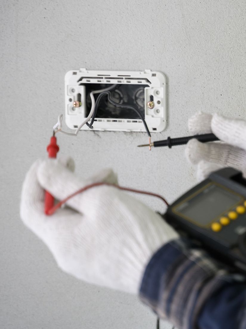 A person wearing white gloves is using a multimeter to test an electrical outlet